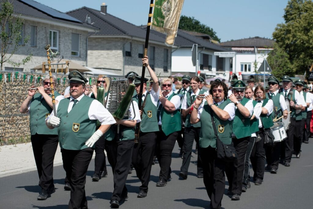 A marching band in military attire with white shirts and green vests. One person is holding up a ornamental flag.