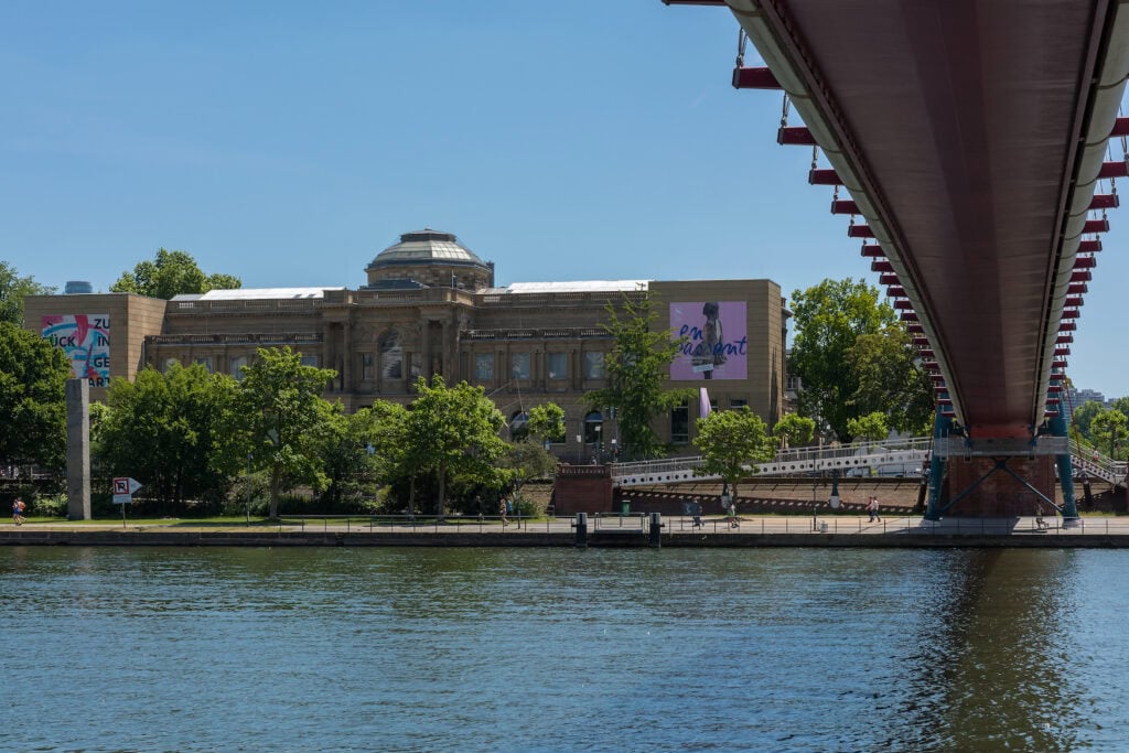 A wide building with two wings and a central dome stands on a riverbank. On the right of the picture is a bridge, photographed from below.