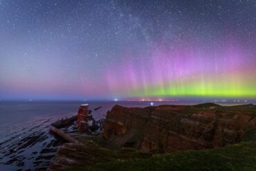 Green and purple auroras shine above the cliffs of the island of Heligoland, with Lange Anna on the left.