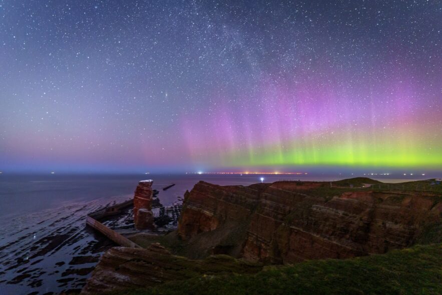 Green and purple auroras shine above the cliffs of the island of Heligoland, with Lange Anna on the left.