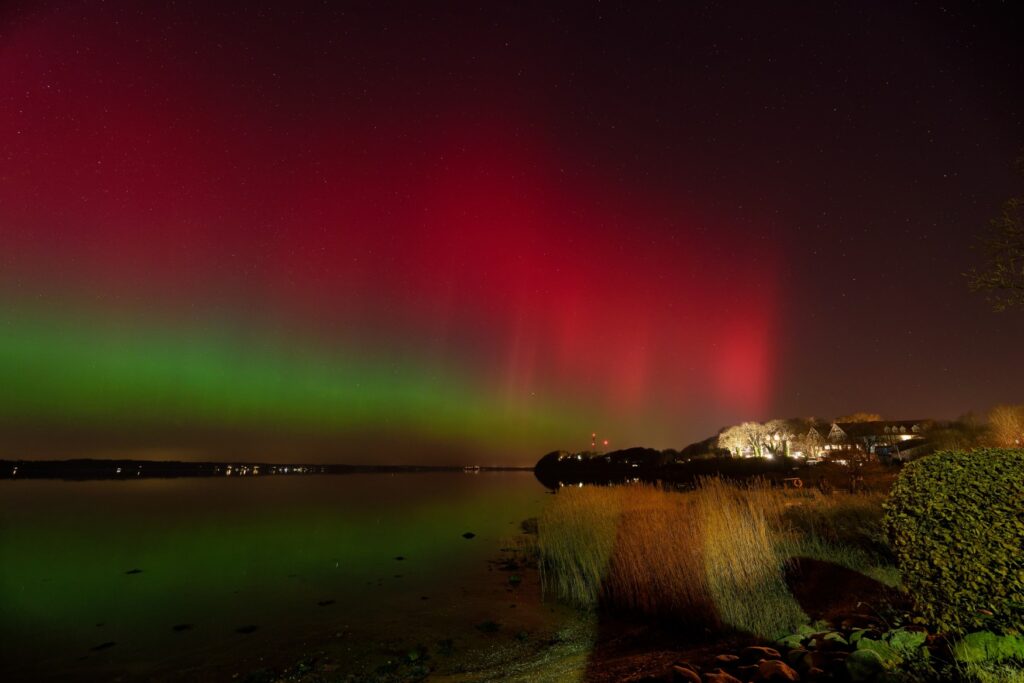 Above a bay, with a few illuminated houses on the shore to the left, blurry red and green auroras shine in the night sky.