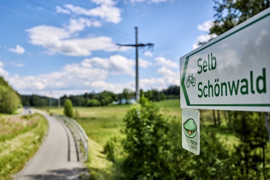 Auf einem weißen Schild weist in grüner Schrift ein Pfeil Richtung Selb und Schönwald, daneben ein Fahrradsymbol. Im Hintergrund unscharf zu erkennen: ein leicht gebogener Aspahltweg, rechts davon ein grünes Feld, im Hintergrund Wald.