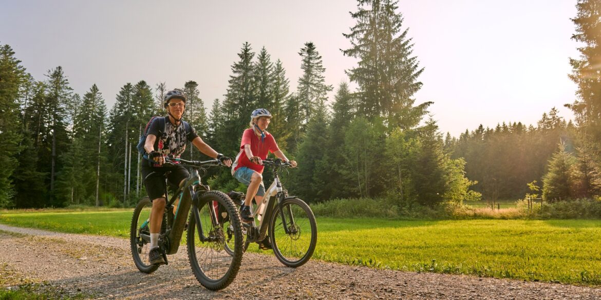 Zwei Frauen in Sportkleidung fahren auf E-Bikes über einen Schotterweg, im Hintergrund Wald. Darüber blauer Himmel mit Sonnenschein.