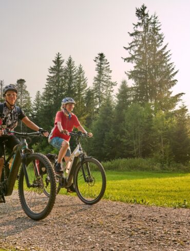 Zwei Frauen in Sportkleidung fahren auf E-Bikes über einen Schotterweg, im Hintergrund Wald. Darüber blauer Himmel mit Sonnenschein.