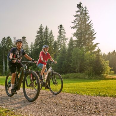Zwei Frauen in Sportkleidung fahren auf E-Bikes über einen Schotterweg, im Hintergrund Wald. Darüber blauer Himmel mit Sonnenschein.