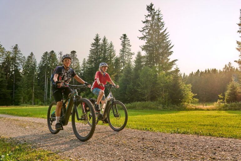 Zwei Frauen in Sportkleidung fahren auf E-Bikes über einen Schotterweg, im Hintergrund Wald. Darüber blauer Himmel mit Sonnenschein.