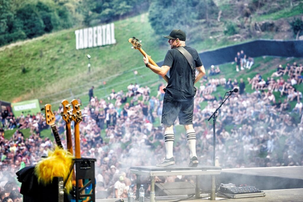 A bass player in skate attire jumping on a festival stage in front of a huge crowd.