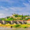 Blick über den Main in Würzburg, mittig eine Brücke, hoch oben auf der anderen Uferseite die mächige Festung der Stadt