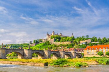 Blick über den Main in Würzburg, mittig eine Brücke, hoch oben auf der anderen Uferseite die mächige Festung der Stadt