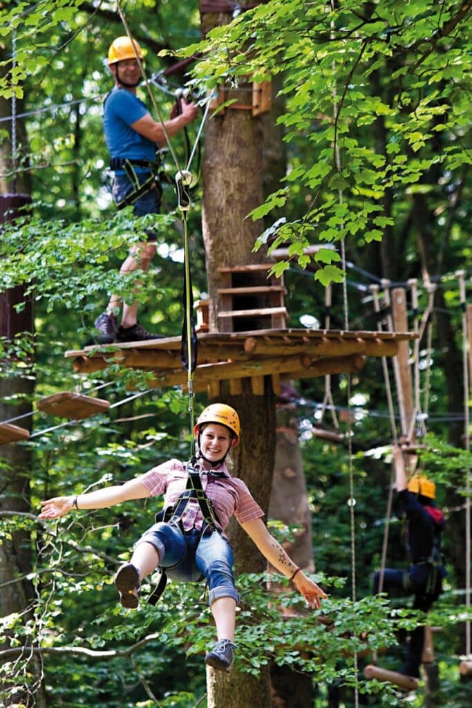 Eine Frau mit gelben Helm und Sicherheitsgurt hängt in einem Kletterwald frei in der Luft und lacht in die Kamera. Auf dem Podest am Baum hinter ihr hängt sich ein Mann, ebenfalls mit Helm, in die Sicherung ein.