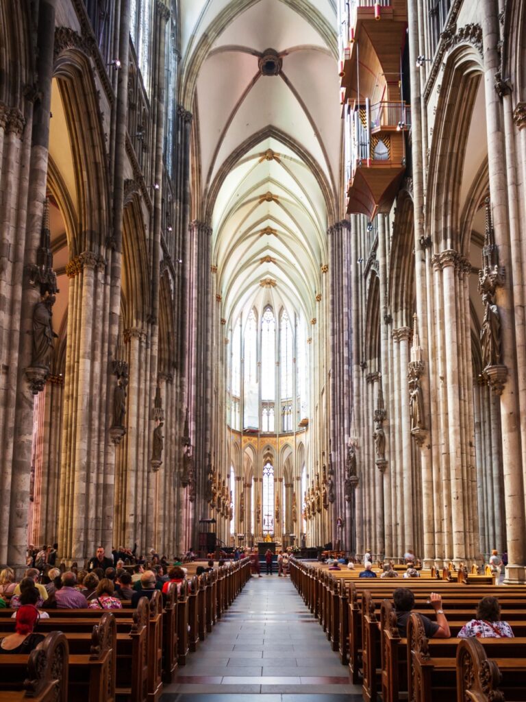 A view of the interior of Cologne Cathedral, looking toward the choir. A few visitors are seated in the pews on either side of the central aisle.