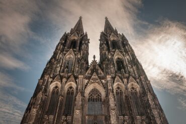 A view of the sunlit west facade of Cologne Cathedral, with its two distinctive towers rising into the blue sky.