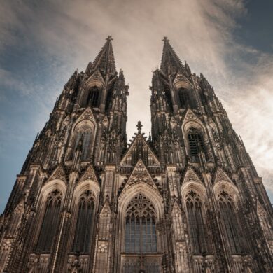 A view of the sunlit west facade of Cologne Cathedral, with its two distinctive towers rising into the blue sky.