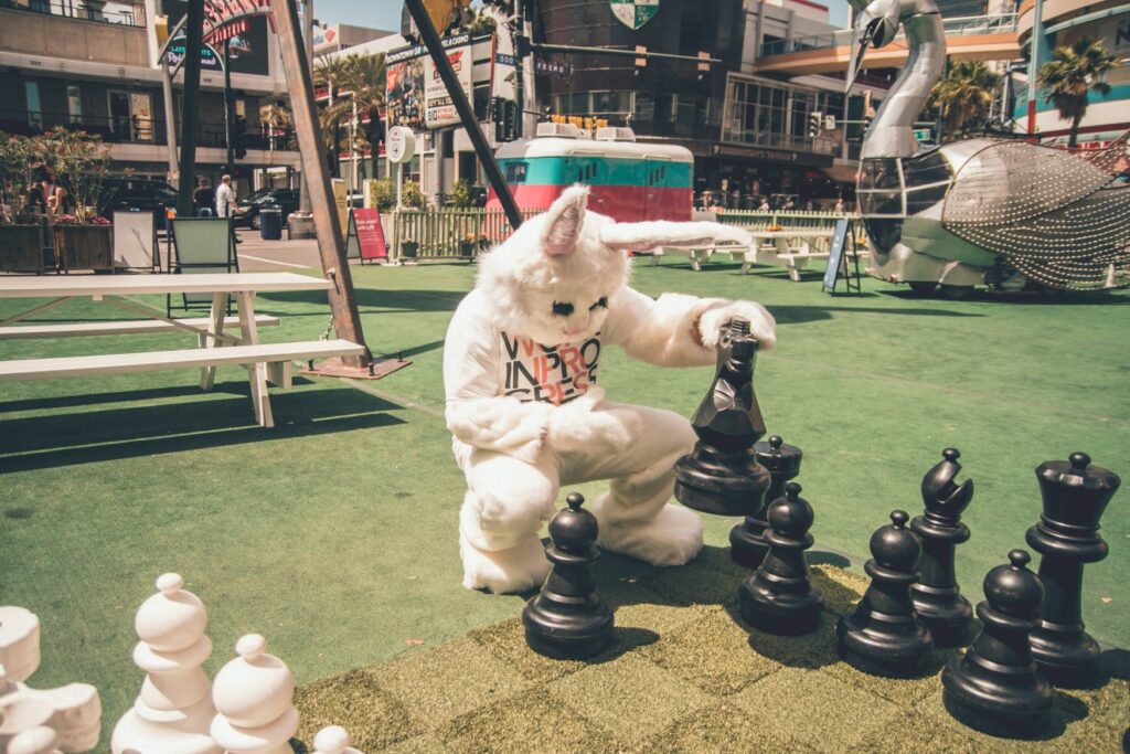 A person in a bunny costume is looking for eggs under an oversized chess piece.