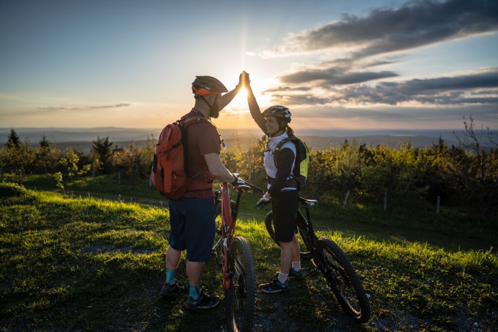 Ein Mann und eine Frau in Sportkleidung und mit Helmen schlagen über ihren Köpfen ein High Five, zwischen ihnen stehen ihre Fahrräder. Sie stehen auf einer grünen Anhöhe, im Hintergrund geht über einem Tal die Sonne unter.