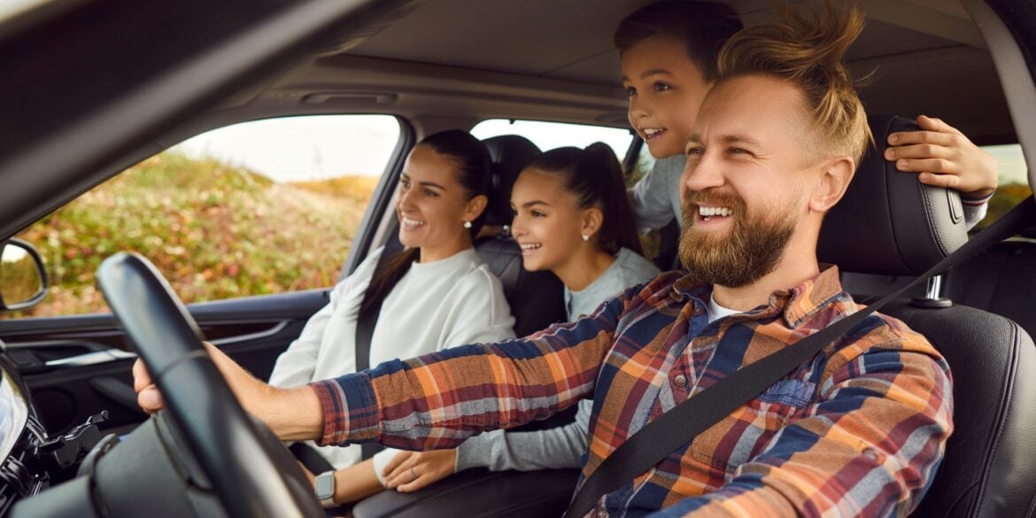 Ein Mann, eine Frau und zwei Kinder sitzen in einem Auto, der Mann am Steuer. Sie haben sichtlich Spaß, dem Mann werden die Haare durch den Wind durchs heruntergelassene Fenster durchgewirbelt.