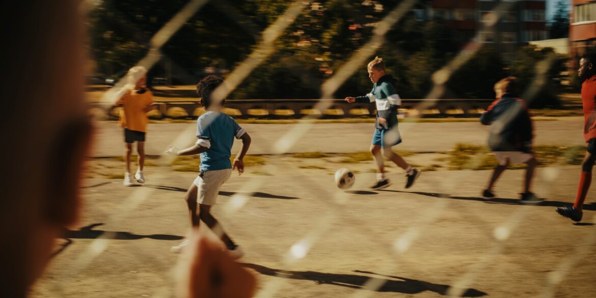 Ein Kind beobachtet durch einen Metallzaun andere Kinder, die auf einem Asphaltplatz Fußball spielen.