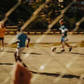 Ein Kind beobachtet durch einen Metallzaun andere Kinder, die auf einem Asphaltplatz Fußball spielen.