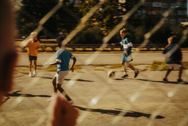 Ein Kind beobachtet durch einen Metallzaun andere Kinder, die auf einem Asphaltplatz Fußball spielen.