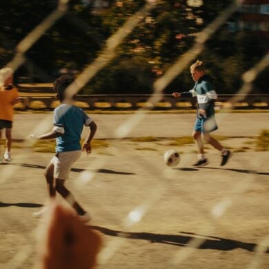 Ein Kind beobachtet durch einen Metallzaun andere Kinder, die auf einem Asphaltplatz Fußball spielen.