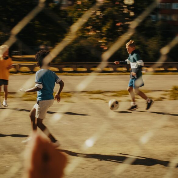 Ein Kind beobachtet durch einen Metallzaun andere Kinder, die auf einem Asphaltplatz Fußball spielen.