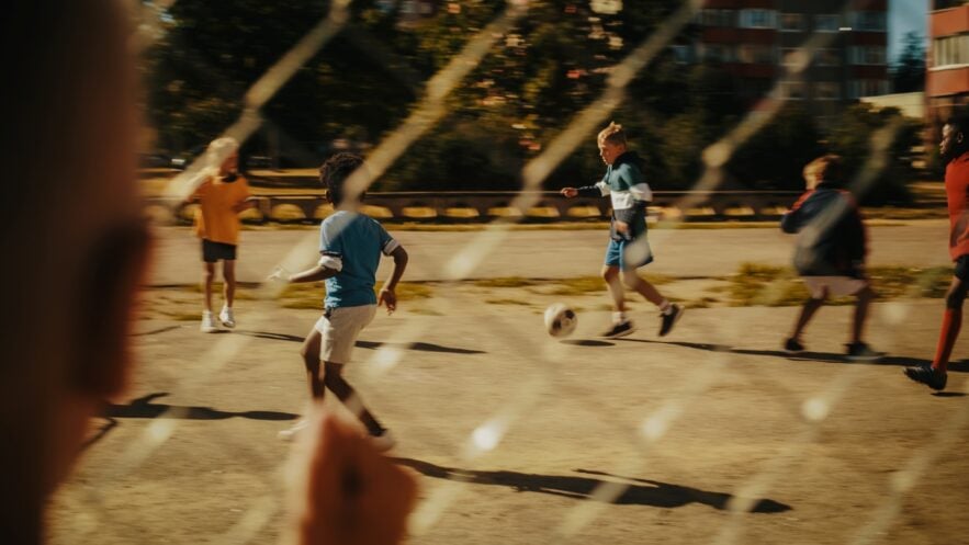 Ein Kind beobachtet durch einen Metallzaun andere Kinder, die auf einem Asphaltplatz Fußball spielen.