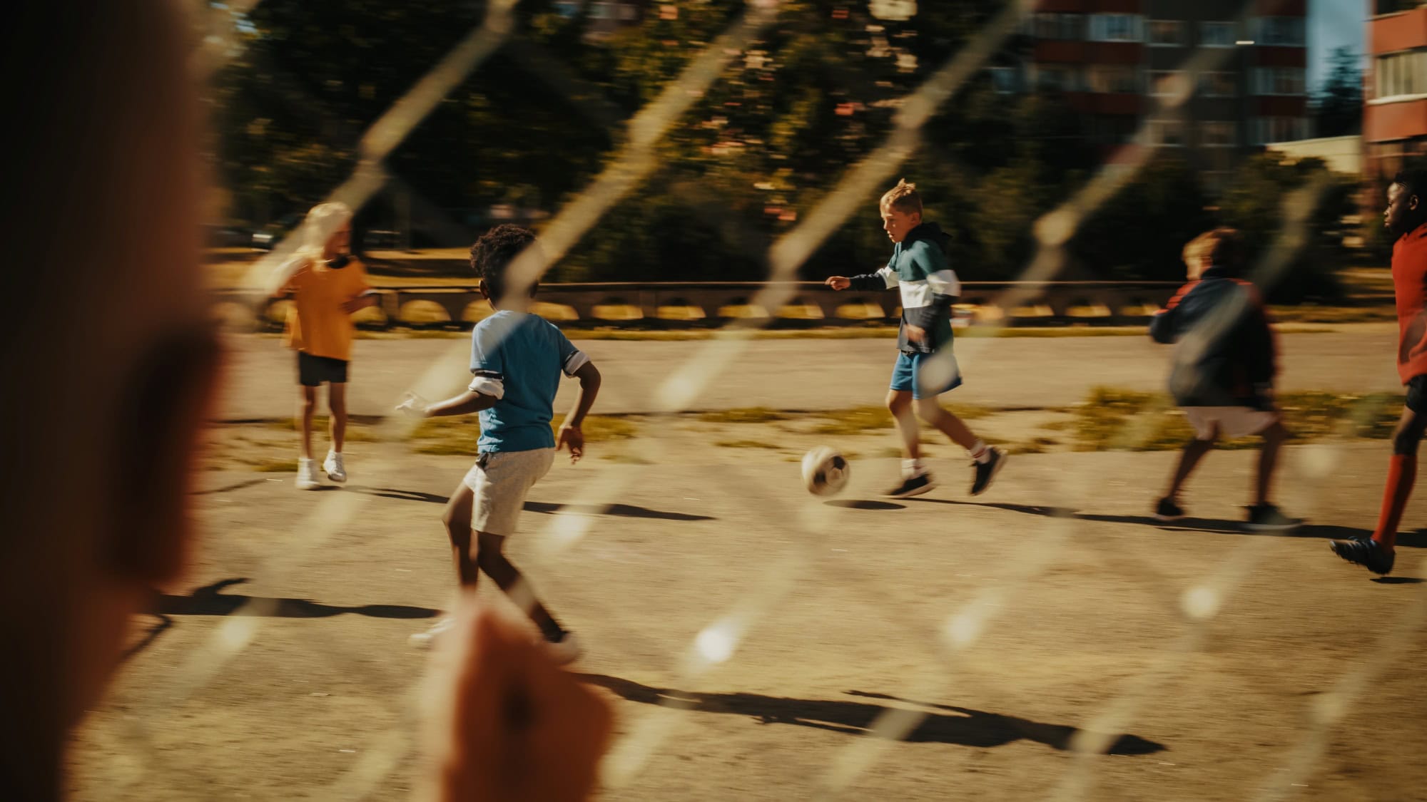 Ein Kind beobachtet durch einen Metallzaun andere Kinder, die auf einem Asphaltplatz Fußball spielen.