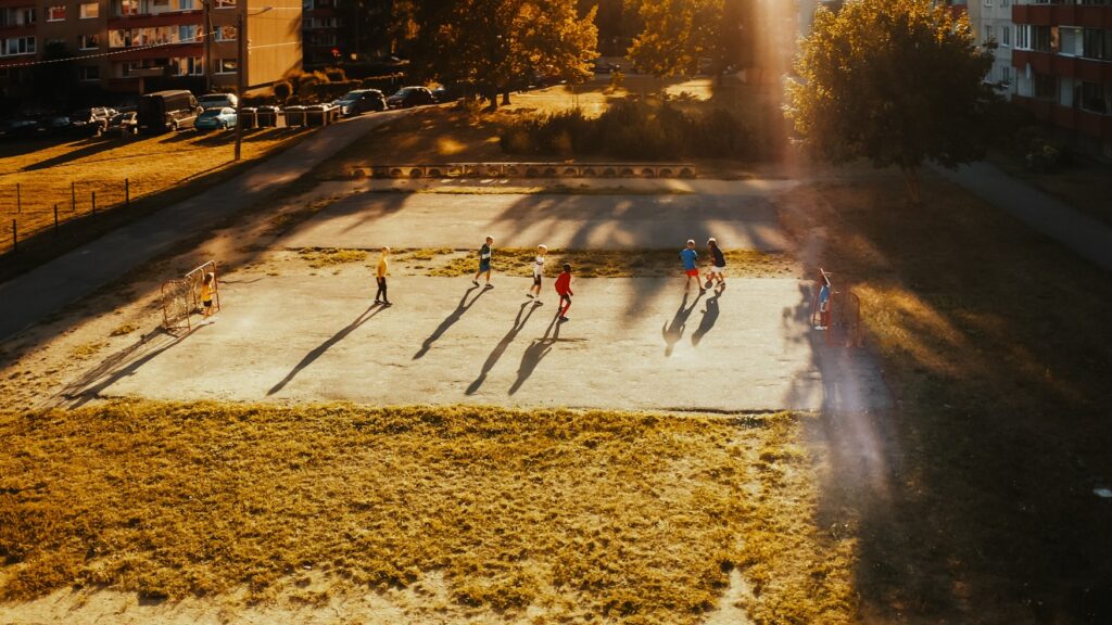 Mehrere Kinder spielen an einem Sommerabend auf einem Platz in Nachbarschaft Fußball. Durch den Sonnenuntergang werfen sie lange Schatten.