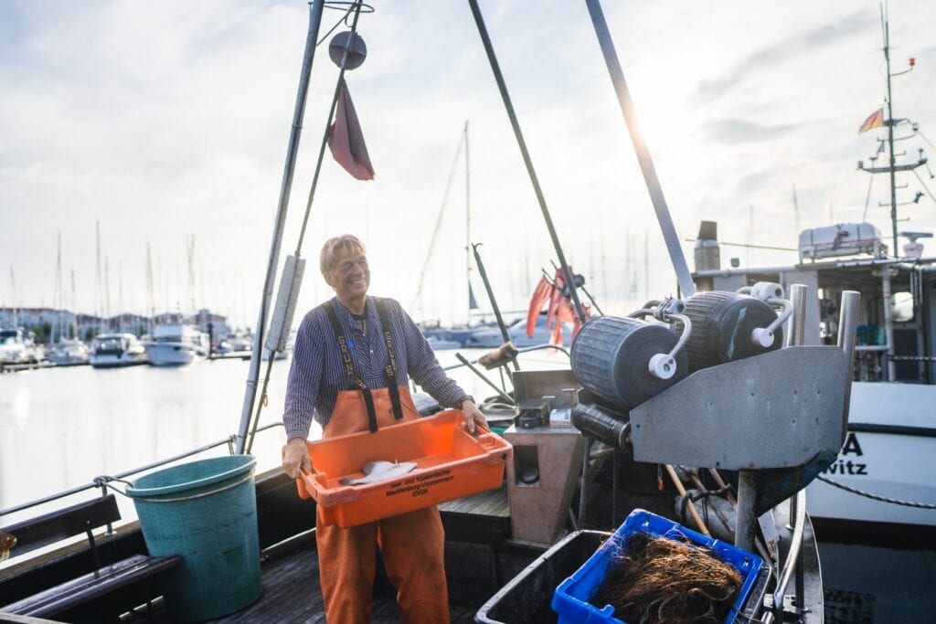 Ein Fischer mit blau-weißem Hemd und orangener Schutzhose steht auf einem Kutter und hält eine orangene Kiste mit seinem Fang in Händen.