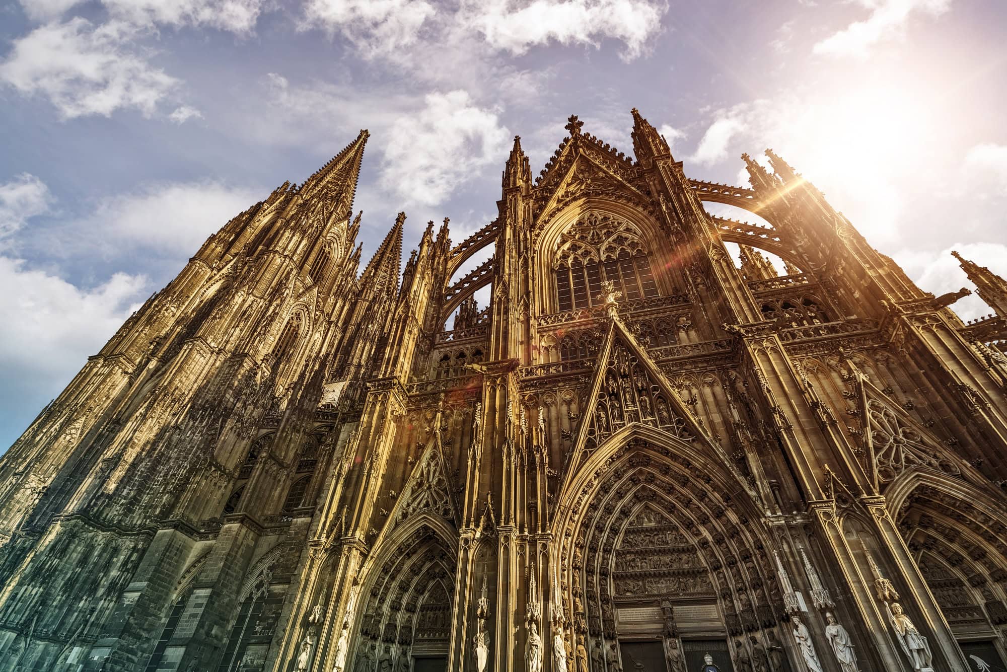 Außenansicht des Kölner Doms, hinter dessen Dach die Sonne hervorscheint.
