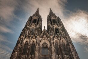 Blick auf die sonnenbeschienene Westfassade des Kölner Doms mit seinen zwei charakteristischen Türmen, die in den blauen Himmel ragen.
