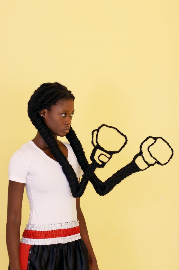 Eine junge, schwarze Frau mit langen, schwarzen Haaren, die in zwei Zöpfe gebunden sind. Die Zöpfe sind am Ende wie zwei Boxhandschuhe geformt, die zum Kampf gehoben sind.