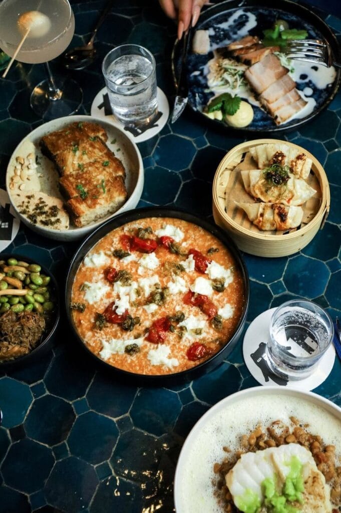 Auf einem gefliesten Tisch stehen mehrere kleine Teller mit unterschiedlichen Gerichten, unter anderem asiatische Teigtaschen und gegrillter Fisch