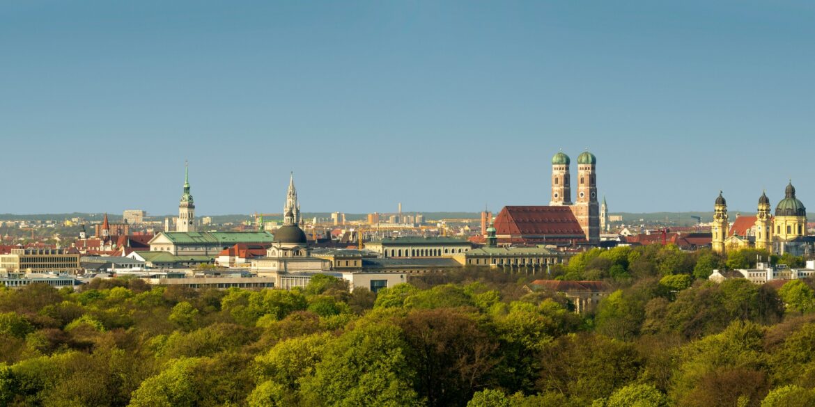 Blick über die Dächer Münchens mit vielen grünen Bäumen und ins sanfte Licht der untergehenden Frühlingssonne getaucht. Im Fokus die charakteristischen Türme der Frauenkirche.