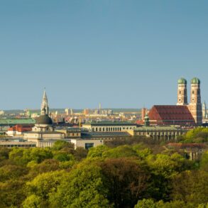 Blick über die Dächer Münchens mit vielen grünen Bäumen und ins sanfte Licht der untergehenden Frühlingssonne getaucht. Im Fokus die charakteristischen Türme der Frauenkirche.