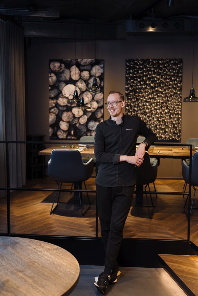 A young man wearing glasses stands in a modern restaurant, dressed in a black chef's uniform, and is laughing. The name “Kevin Romes” is visible on his jacket.