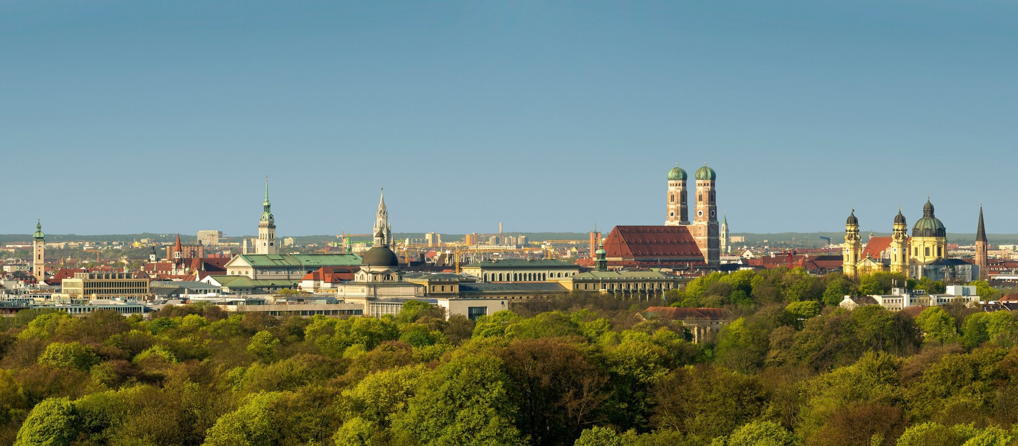A view over the rooftops of Munich, dotted with green trees and bathed in the soft light of the setting spring sun.