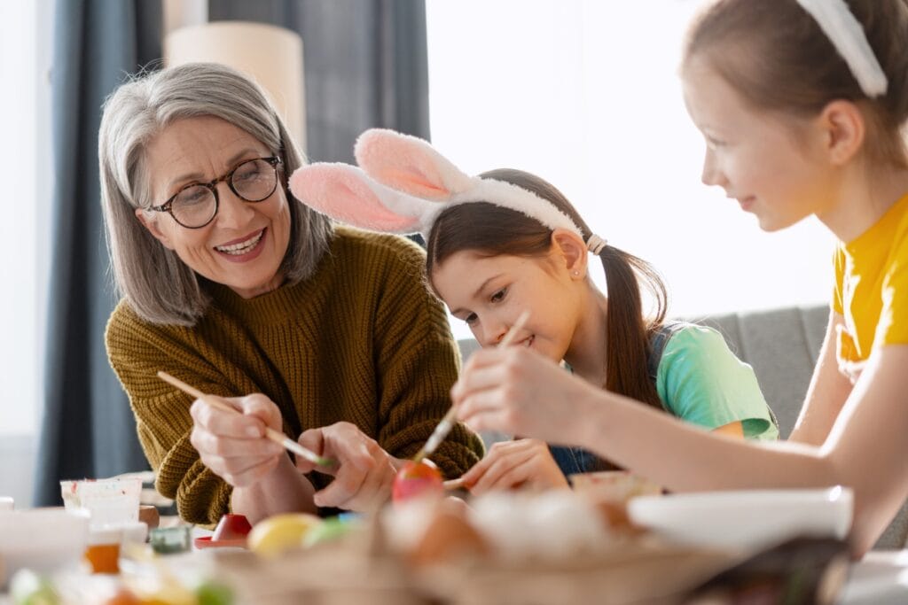 Zwei Kinder bemalen mit ihrer Großmutter Ostereier. Die Kinder haben dabei flauschige Hasenohren angezogen.