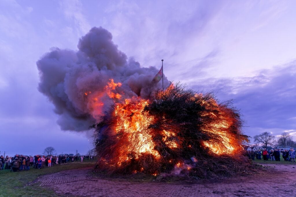 Auf einem steinernen Platz brennt ein großes, rundes Osterfeuer. Im Halbkreis stehen Menschen darum.