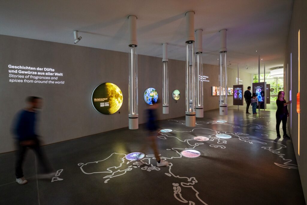 In a museum, a walk-on world map is painted on the floor. Several locations are marked, and on the wall next to it are explanations of specific scents and spices that originally come from those marked locations.
