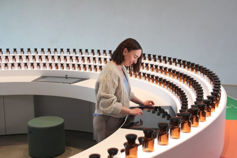 Eine junge Frau bedient einen in eine halbrunde Anrichte eingelassenen Touchscreen. Am äußeren Rand der Anrichte sind eine Vielzahl kleiner Apothekerfläschchen im Halbrund aufgestellt.