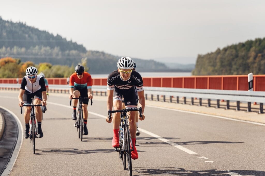 Eine Gruppe Rennradfahrer fährt über eine Brücke in Richtung des Betrachters. Rechts im Hintergrund ist ein See oder ein Fluss zu erkennen, links zieht sich ein von Wald bewachsener Hang zum oberen Bildrand.