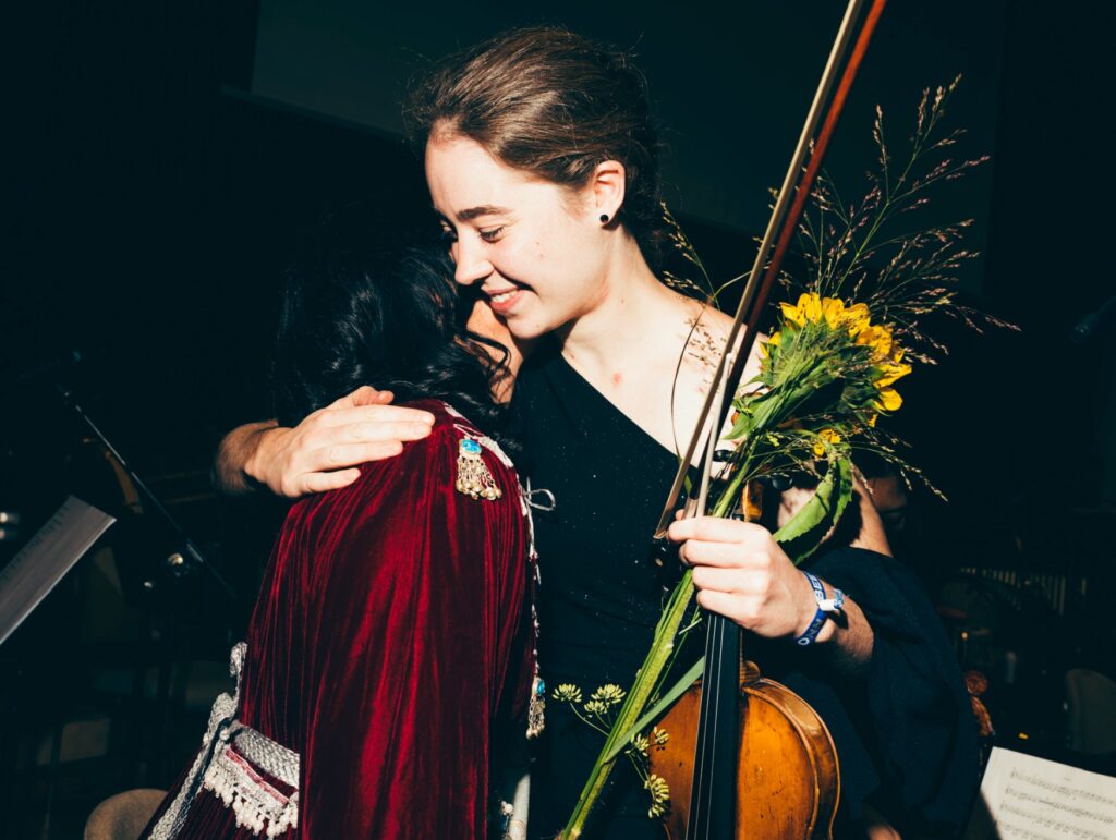 Two young women, one of them holding a violin, embrace after a concert.