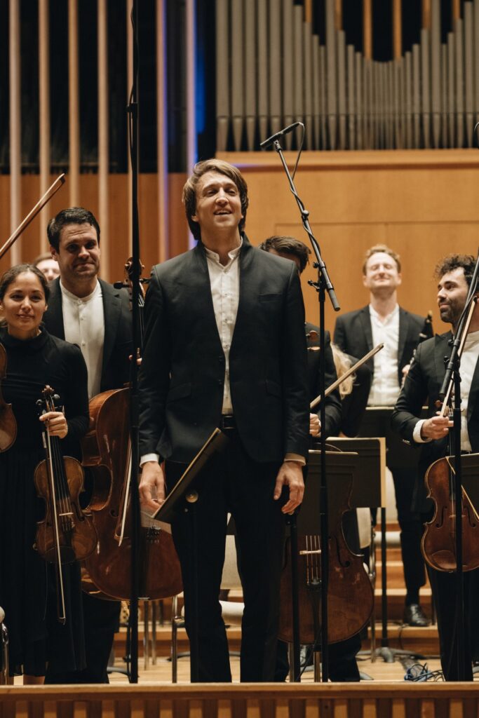 Ein Dirigent steht vor seinem Orchester und lächelt ins Publikum.