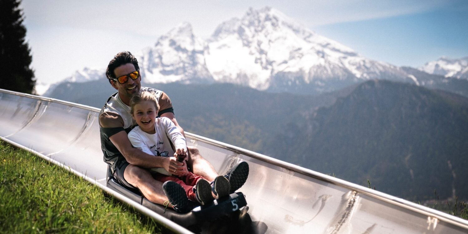 Ein Mann und ein Kinder fahren auf einer Sommerrodelbahn einen Hang hinab. Im Hintergrund sieht man einen schneebedeckten Gipfel der Alpen.