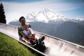 Ein Mann und ein Kinder fahren auf einer Sommerrodelbahn einen Hang hinab. Im Hintergrund sieht man einen schneebedeckten Gipfel der Alpen.