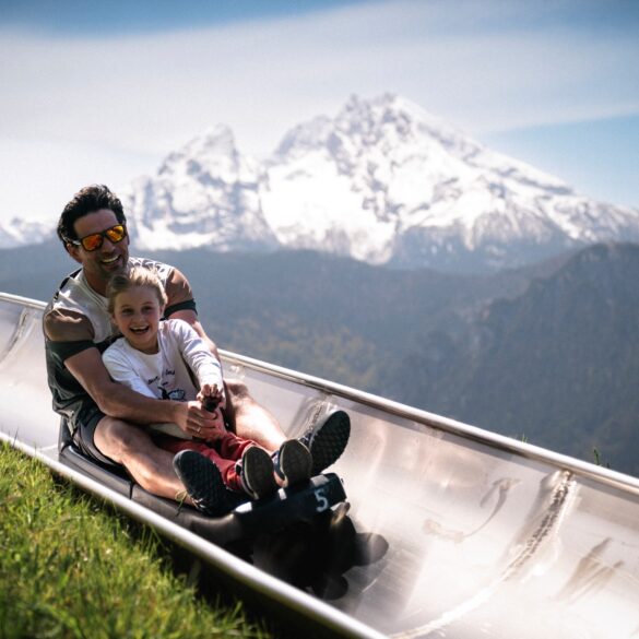 Ein Mann und ein Kinder fahren auf einer Sommerrodelbahn einen Hang hinab. Im Hintergrund sieht man einen schneebedeckten Gipfel der Alpen.