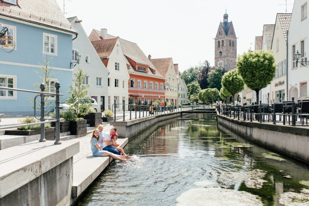 Eine Familie mit zwei kleinen Kindern hält in einer sommerlichen Kleinstadt mit hübschen, historischen Häusern die Füße in einen kanaliserten Bach. 