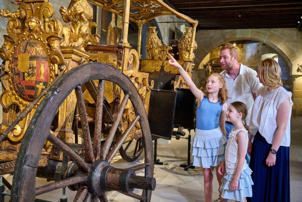 Eine Familie mit zwei kleinen Kindern bestaunt eine goldene Kutsche in einer historischen Ausstellung.