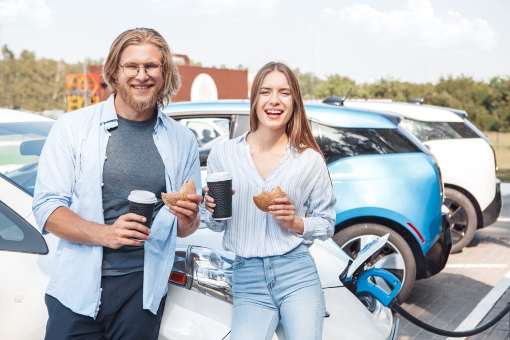 A young man and a woman are standing at a rest stop, leaning against their car, each holding a roll and a cup of coffee. In the background, a charging cable is plugged into the car.